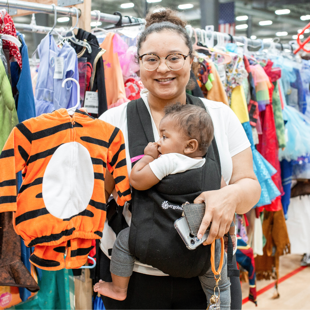 A Mom and her Infant in a Carrier holds up up a Tiger Costume, she is excited to buy.
